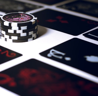 A stack of poker chips with a distinctive black and white pattern is placed on a table covered with several playing cards. The cards are dark with red and white symbols, creating a bold contrast against the lighter surface.