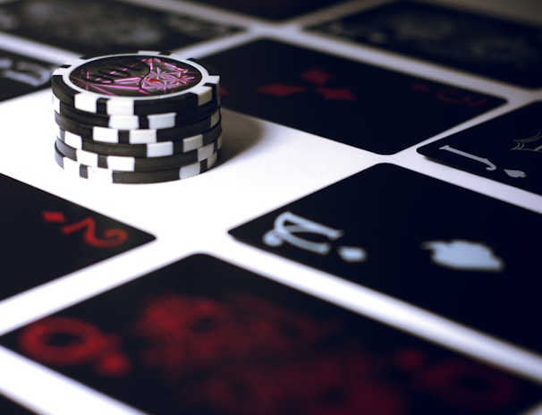 A stack of poker chips with a distinctive black and white pattern is placed on a table covered with several playing cards. The cards are dark with red and white symbols, creating a bold contrast against the lighter surface.