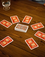 A wooden table with several playing cards arranged in a circle around a stack of cards in the center. Each card has a red back with text and illustrations. Two drinking glasses, one with a clear liquid, are also on the table, with a person's arm visible in the background.