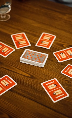 A wooden table with several playing cards arranged in a circle around a stack of cards in the center. Each card has a red back with text and illustrations. Two drinking glasses, one with a clear liquid, are also on the table, with a person's arm visible in the background.