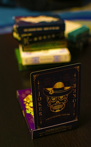 A playing card featuring a stylized sugar skull design, labeled 'Joker', is prominently displayed in the foreground. The card has an intricate design with a hat on the skull and decorative elements in gold. In the background, multiple stacks of board games or boxes are slightly out of focus, placed on a dark tabletop.