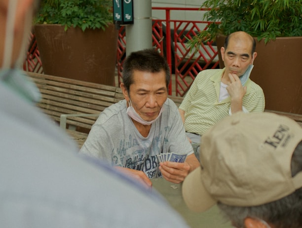 Several individuals gather outdoors, sitting on benches and engaging in a game of cards. The focal point is on a man holding a fan of playing cards, with others observing or participating. Potted plants and a red railing are visible in the background, contributing to the setting's casual and social atmosphere.