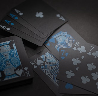 A set of playing cards with a dark and stylish design featuring black backgrounds and white and blue symbols. The cards include a King, Queen, and several numbered cards, all laid out on a dark surface.