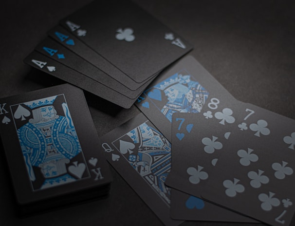 A set of playing cards with a dark and stylish design featuring black backgrounds and white and blue symbols. The cards include a King, Queen, and several numbered cards, all laid out on a dark surface.