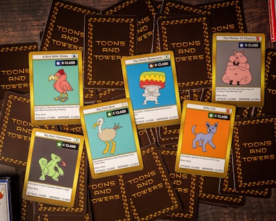 A collection of trading cards from a game called 'Toons and Towers' is spread out on a wooden surface. Each card features a colorful cartoon character and various stats like class, attack, and defense. The characters include a bird, a mushroom, a pig, a chameleon, a cat, and a large bird-like creature.