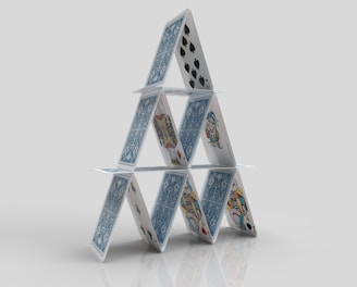 A structure composed of playing cards arranged in a triangular formation, resembling a house of cards. The cards feature various designs and suits, including spades and face cards, standing on a reflective surface against a plain background.