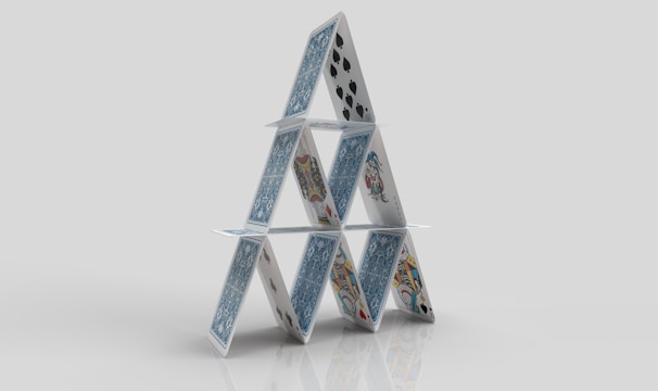 A structure composed of playing cards arranged in a triangular formation, resembling a house of cards. The cards feature various designs and suits, including spades and face cards, standing on a reflective surface against a plain background.