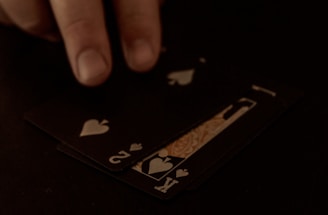 A hand is touching a stack of playing cards laid out on a dark surface. The cards are primarily black, featuring heart suits, including the king and jack. The scene is dimly lit, giving it an intimate or mysterious atmosphere.