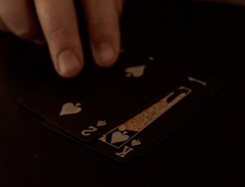 A hand is touching a stack of playing cards laid out on a dark surface. The cards are primarily black, featuring heart suits, including the king and jack. The scene is dimly lit, giving it an intimate or mysterious atmosphere.