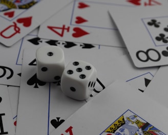 A collection of playing cards spread out, featuring various suits such as hearts, clubs, and spades. Two white dice with black dots are placed prominently on top of the cards.
