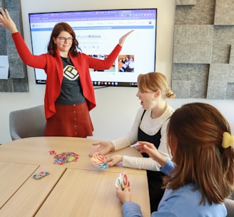 Four people are engaged in a lively game around a wooden table. One person stands with raised arms, while three others are seated, focusing on colorful cards in their hands. There is a computer monitor in the background displaying a webpage, and the room features a modern design with gray acoustic panels on the walls.