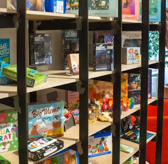 A bookshelf contains a variety of board games, puzzles, and card games. The shelves display colorful and diverse game boxes, with some dedicated to adventure themes. The scene is lively and inviting, with a mix of cartoonish and realistic illustrations on the packaging.