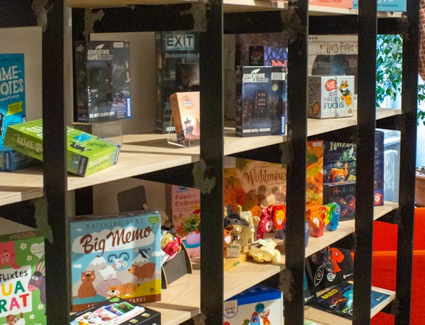 A bookshelf contains a variety of board games, puzzles, and card games. The shelves display colorful and diverse game boxes, with some dedicated to adventure themes. The scene is lively and inviting, with a mix of cartoonish and realistic illustrations on the packaging.