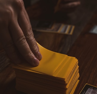 A hand is holding a stack of playing cards with yellow sleeves on a wooden table. Several cards are laid out in the background, and a dice is visible to the side.