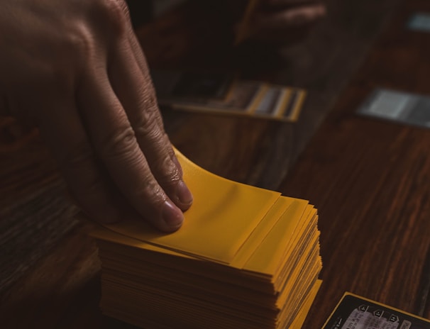 A hand is holding a stack of playing cards with yellow sleeves on a wooden table. Several cards are laid out in the background, and a dice is visible to the side.
