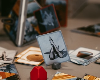 A tabletop game setup with various illustrated cards displayed on a surface. A red, geometric game piece is positioned next to the cards, adding a pop of color. The cards have images and symbols, suggesting a fantasy or strategy theme. The background shows a partially obscured game box with text.
