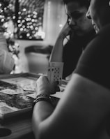 Three people are engaged in a card game, sitting around a table in a cozy, dimly lit setting. One player is holding an Ace and a Ten of diamonds. There are fairy lights and a blurred Christmas tree in the background, adding a festive and warm atmosphere to the scene.